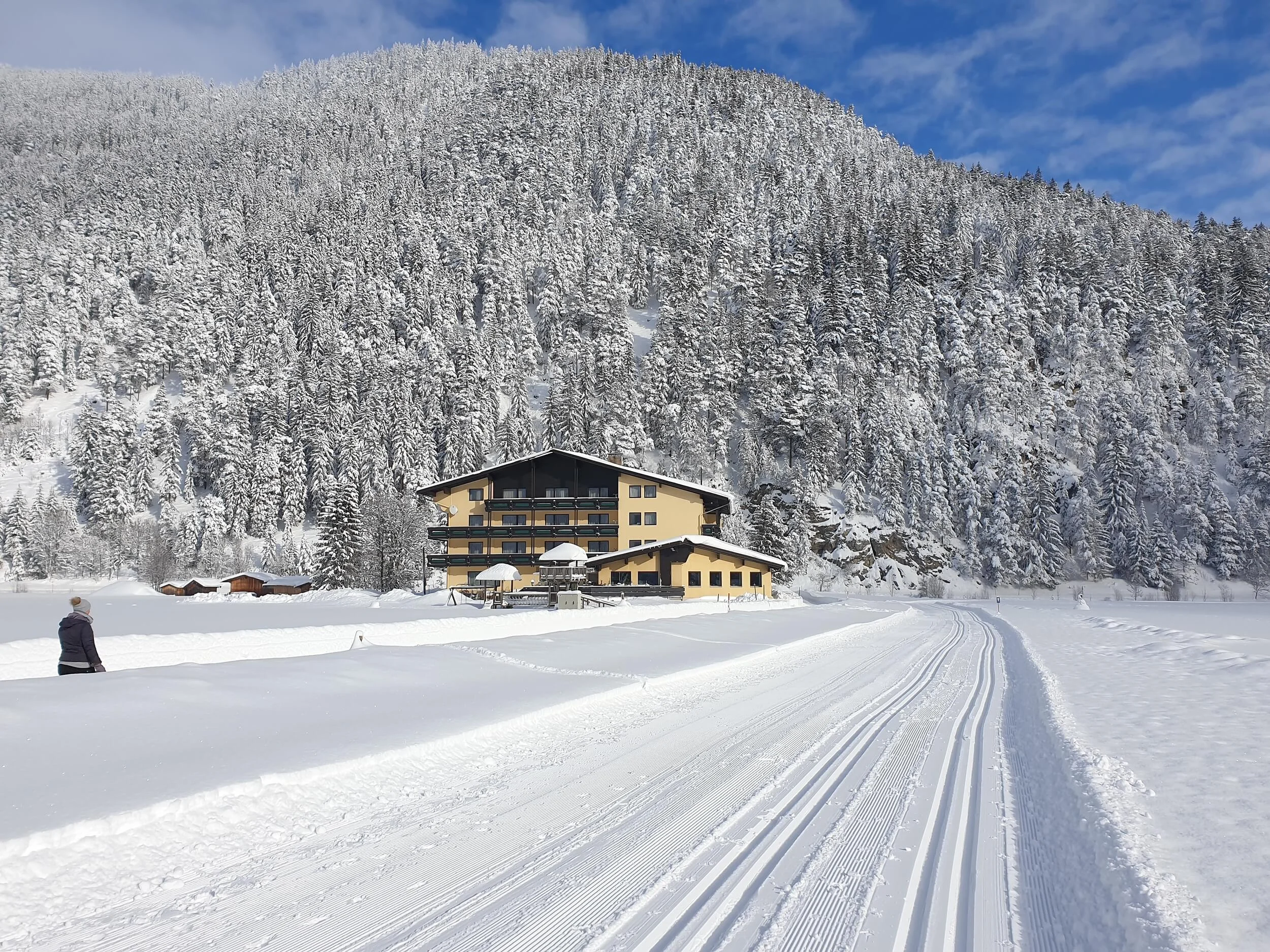 Snow-covered house at the base of a snowy forested hill under blue sky