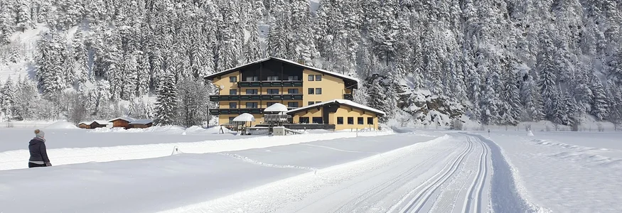 Snow-covered house at the base of a snowy forested hill under blue sky