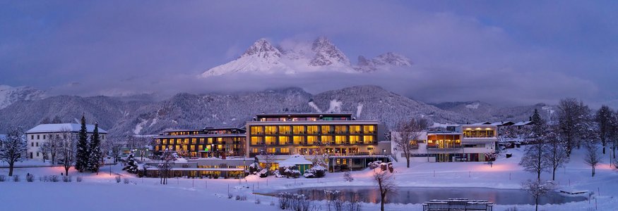 Beleuchtetes Hotel im verschneiten Tal vor bewölktem Berggipfel bei Abenddämmerung