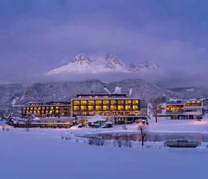 Beleuchtetes Hotel im verschneiten Tal vor bewölktem Berggipfel bei Abenddämmerung