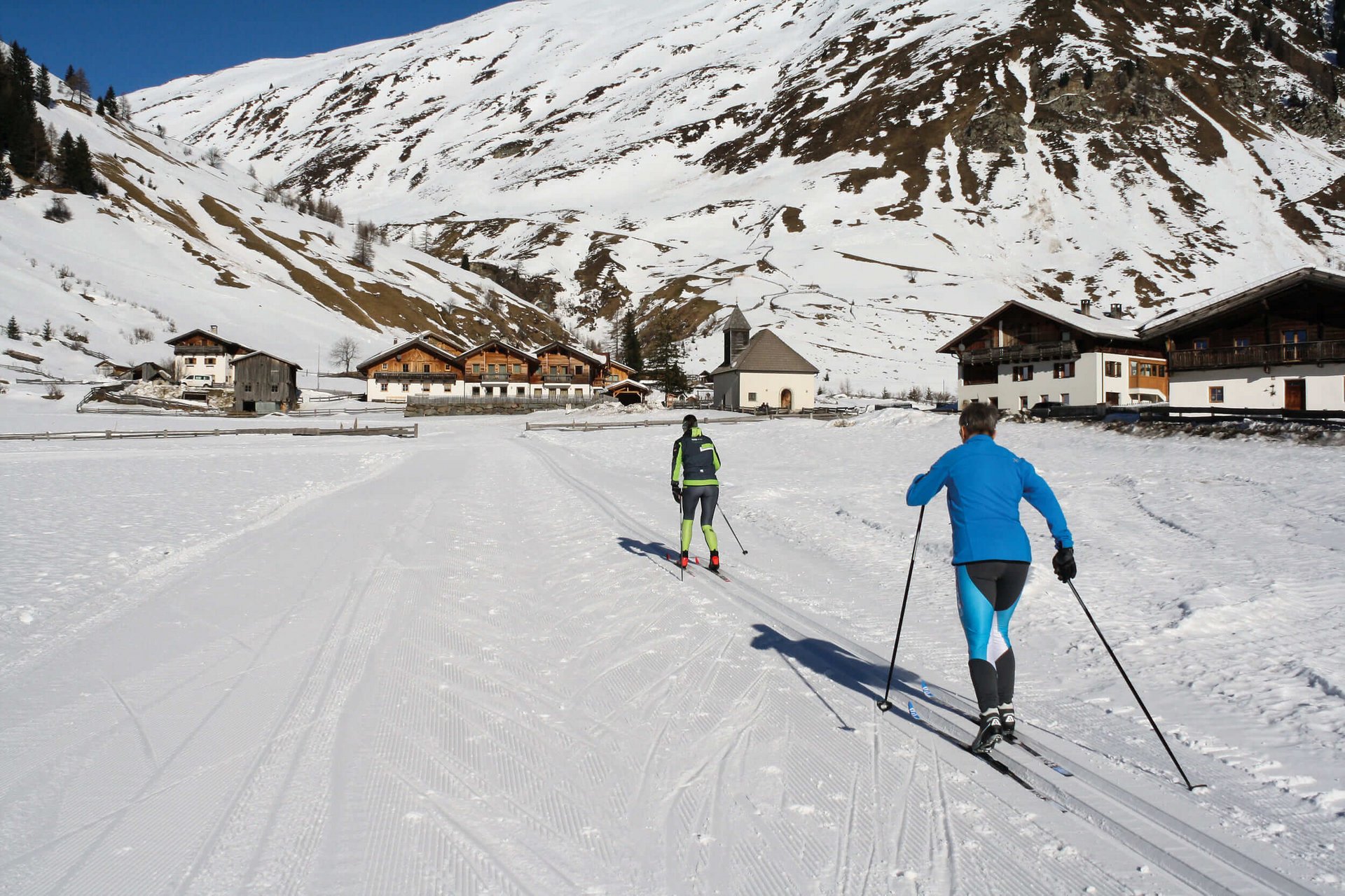 Langlauf Urlaub © Judith Vasselai Zwei Skilangläufer in verschneitem Dorf vor Bergkulisse