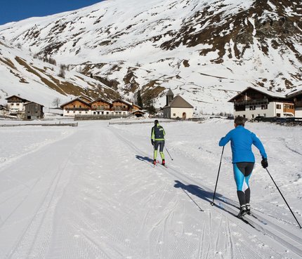 Zwei Skilangläufer in verschneitem Dorf vor Bergkulisse