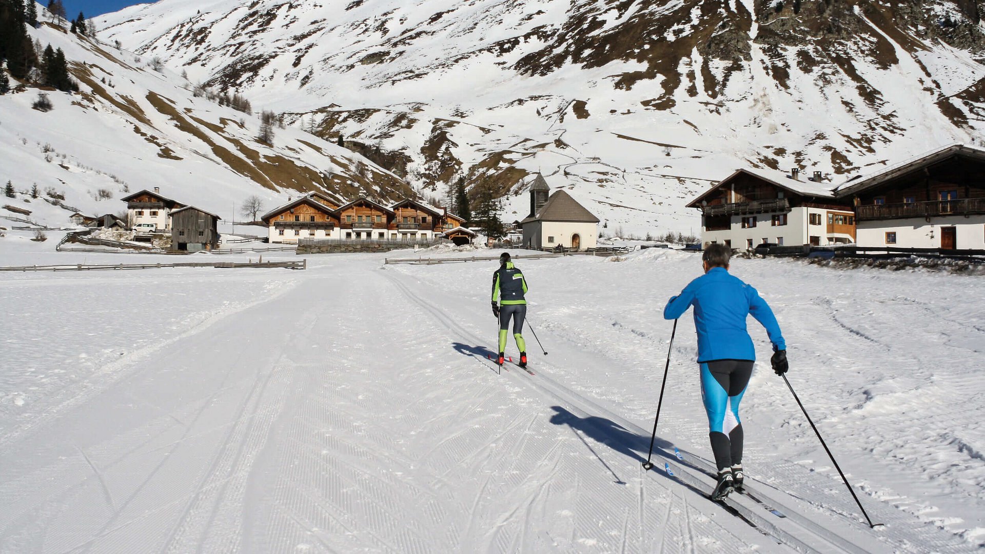 Zwei Skilangläufer in verschneitem Dorf vor Bergkulisse