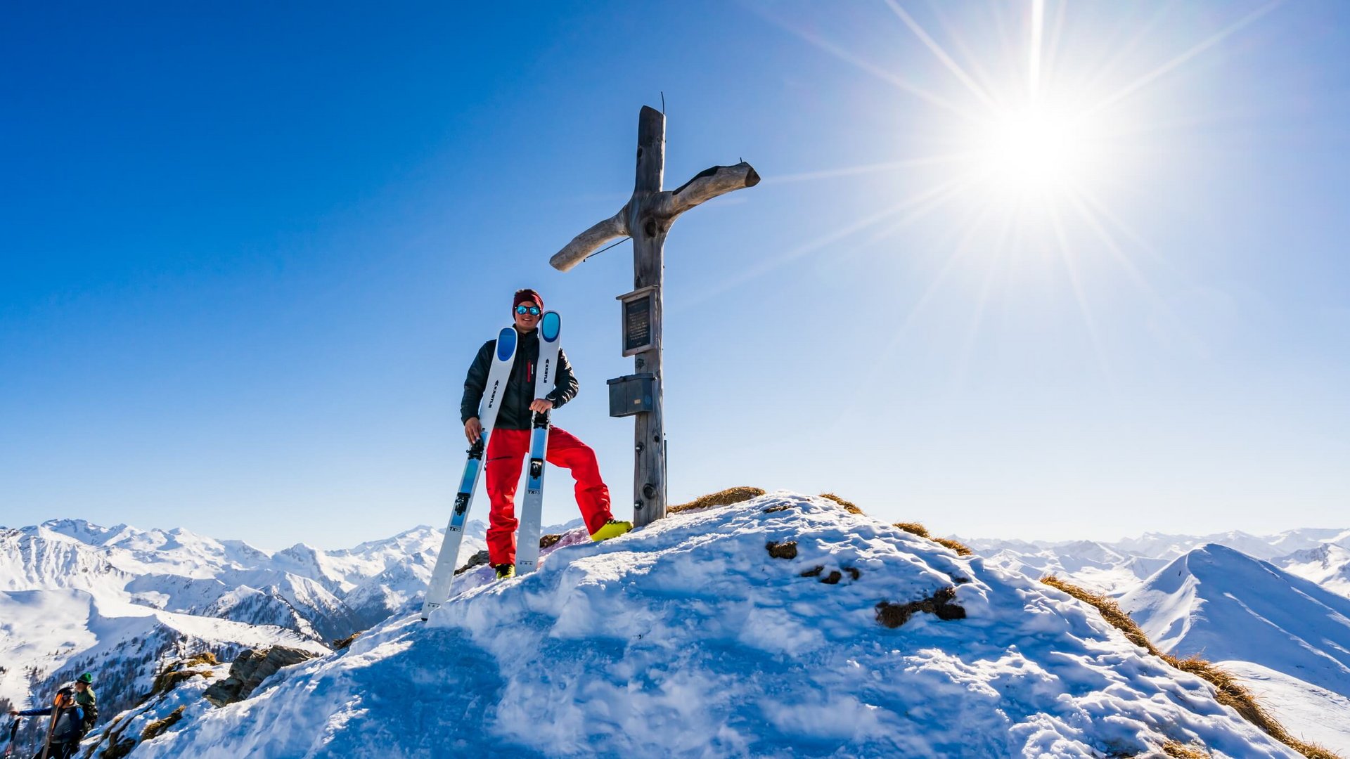 Skifahrer steht an einem Gipfelkreuz auf schneebedecktem Berg bei Sonnenschein