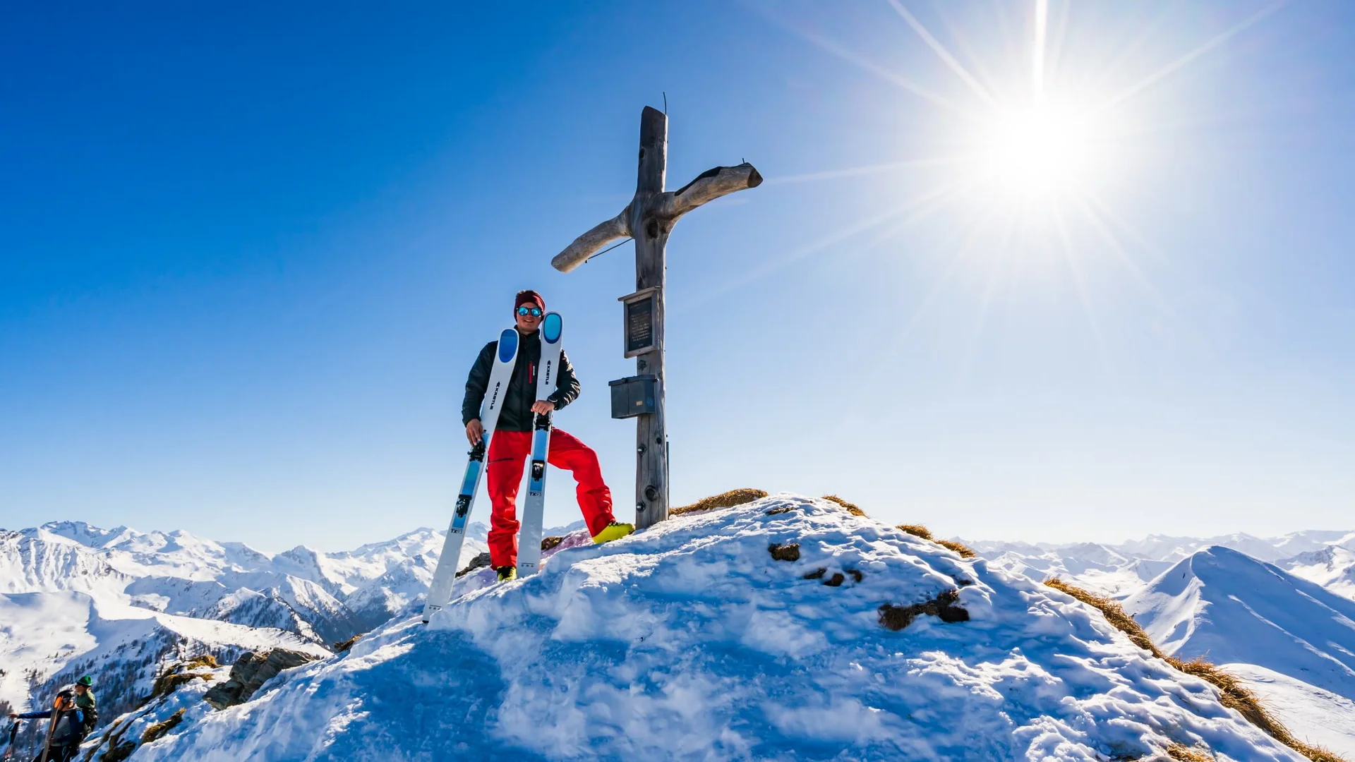 Skifahrer steht an einem Gipfelkreuz auf schneebedecktem Berg bei Sonnenschein