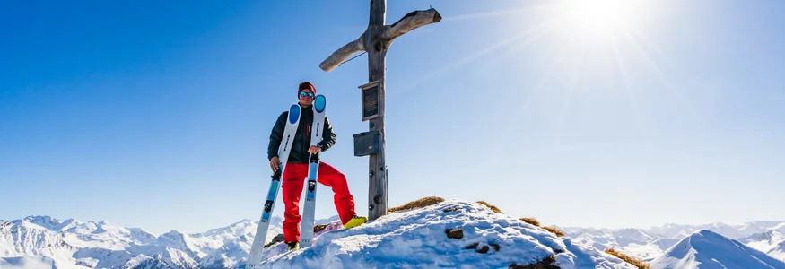 Skifahrer steht an einem Gipfelkreuz auf schneebedecktem Berg bei Sonnenschein