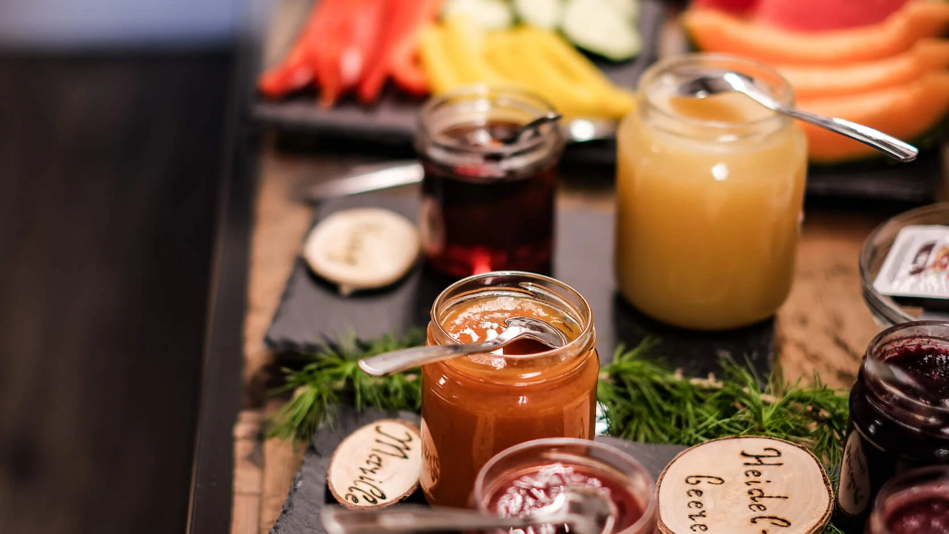 Various jars of jam with cold cuts and vegetables in the background