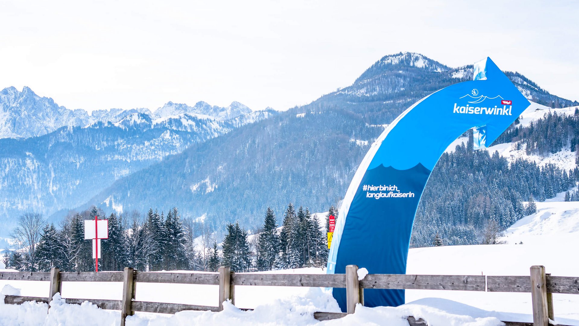 Blauer Pfeil mit Kaiserwinkl-Logo im verschneiten alpinen Winterlandschaft