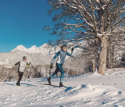 Zwei Personen beim Langlaufen vor verschneiten Bergen und klarem Himmel