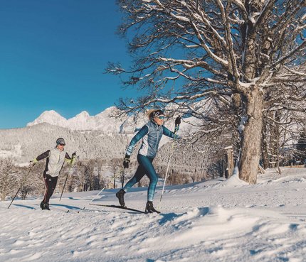 Zwei Personen beim Langlaufen vor verschneiten Bergen und klarem Himmel