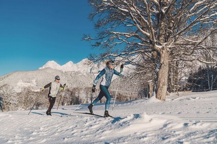 Zwei Personen beim Langlaufen vor verschneiten Bergen und klarem Himmel
