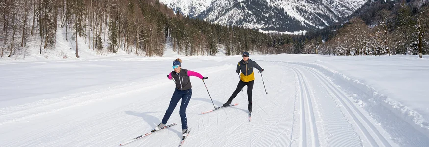 Zwei Langläufer auf verschneiter Loipe vor Bergkulisse