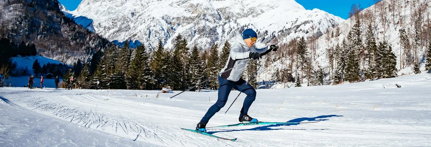 Person beim Langlauf vor verschneiten Bergen und blauem Himmel
