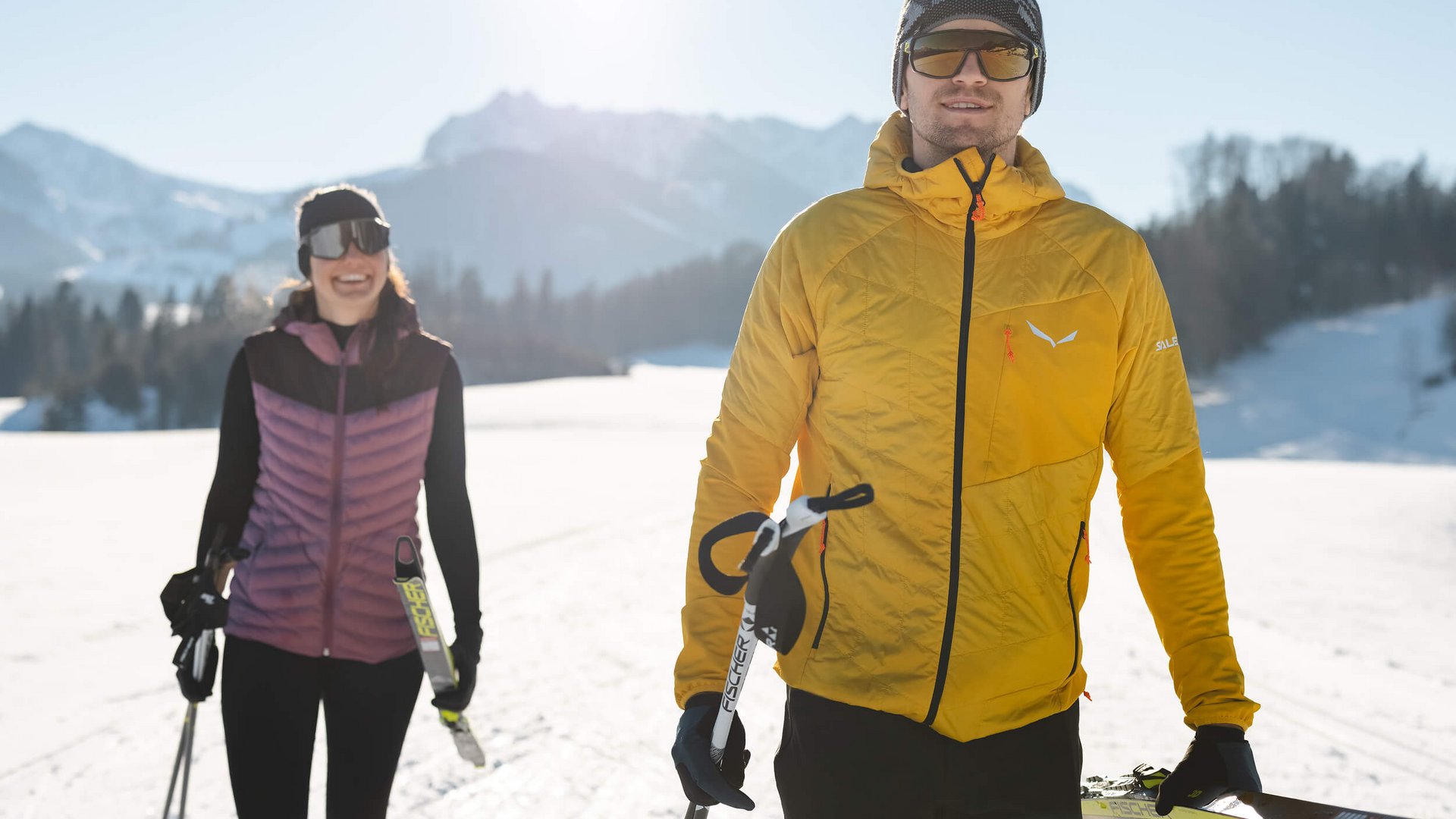 Two cross-country skiers holding equipment in sunny snowy mountain landscape