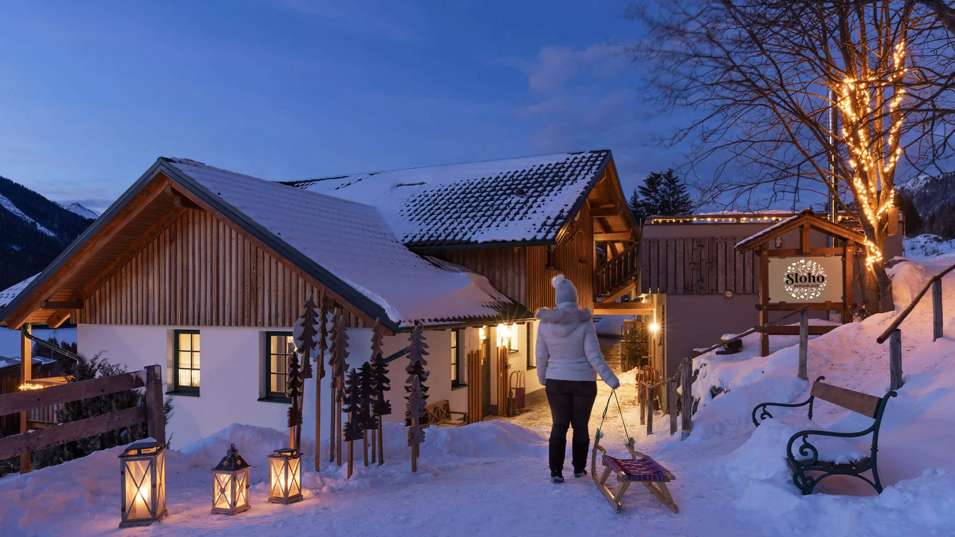 SLOHO Bergurlaub Apartments © Cathrine Stukhard Verschneite Chalets mit Laternen, einer gehenden Person und beleuchtetem Baum