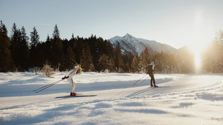 Two cross-country skiers in snow with forest and mountains in sunlight