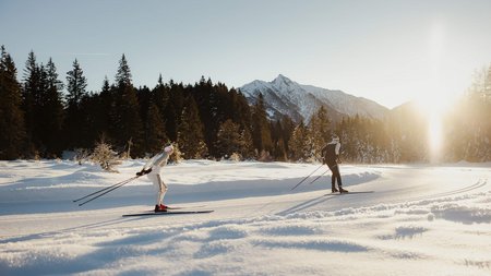 Zwei Langläufer im Schnee vor bewaldeten Bergen bei Sonnenschein