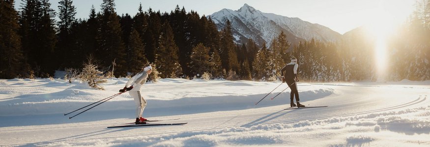 Two cross-country skiers in snow with forest and mountains in sunlight
