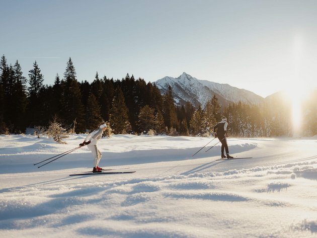 Zwei Langläufer im Schnee vor bewaldeten Bergen bei Sonnenschein