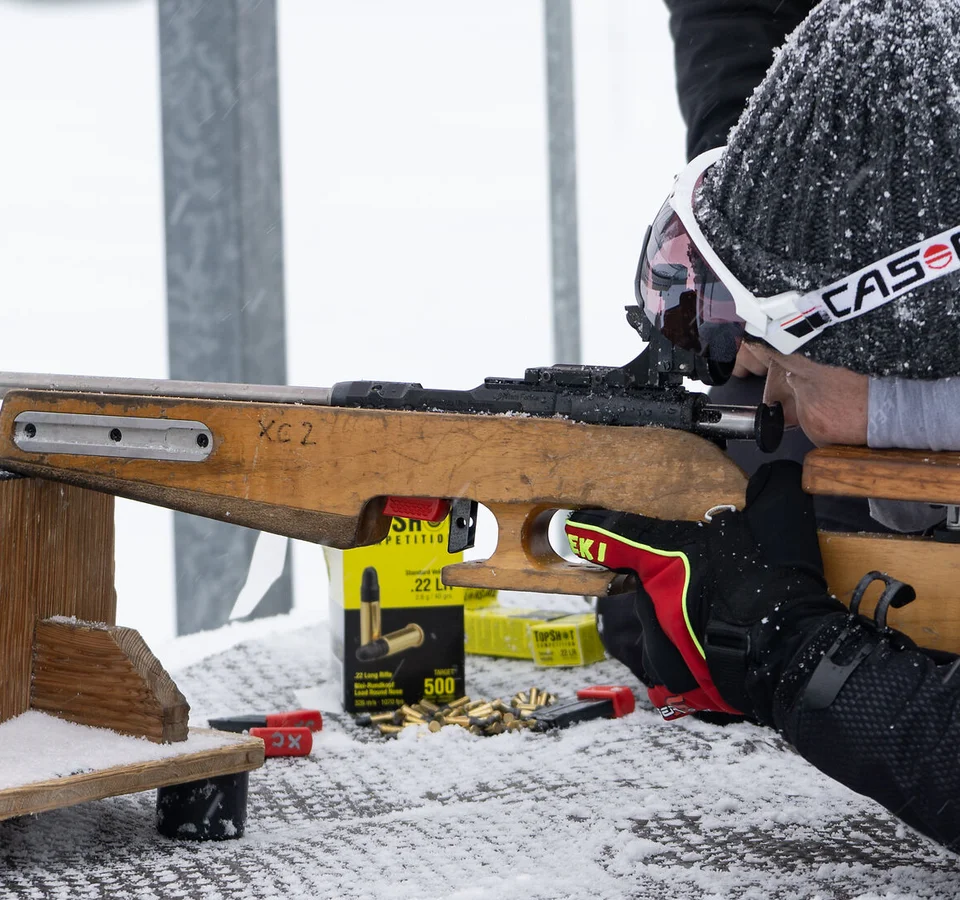 Person im Schnee zielt mit Gewehr auf einem Holztisch in liegender Position