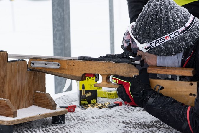 Person im Schnee zielt mit Gewehr auf einem Holztisch in liegender Position