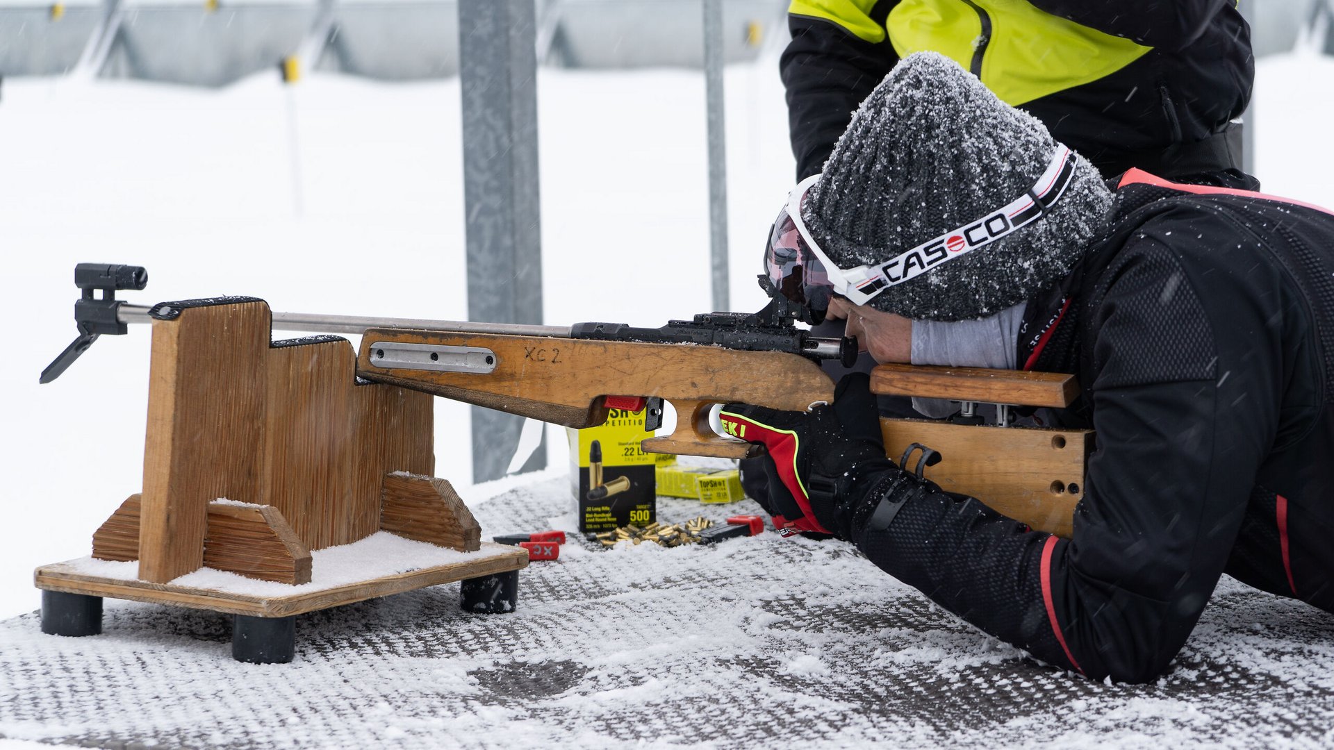Person im Schnee zielt mit Gewehr auf einem Holztisch in liegender Position