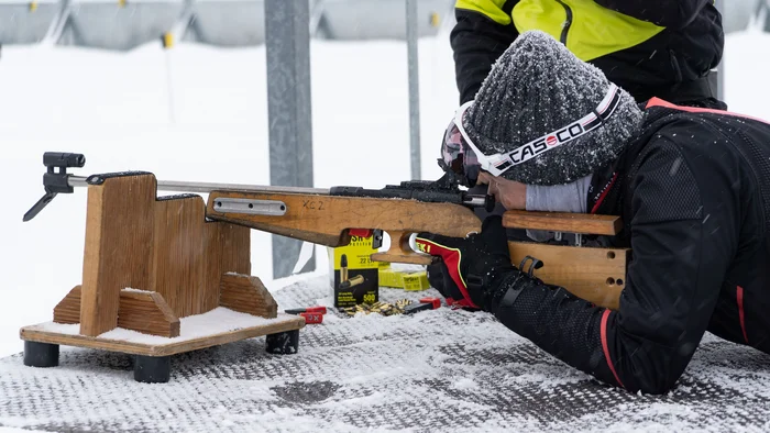 Person im Schnee zielt mit Gewehr auf einem Holztisch in liegender Position