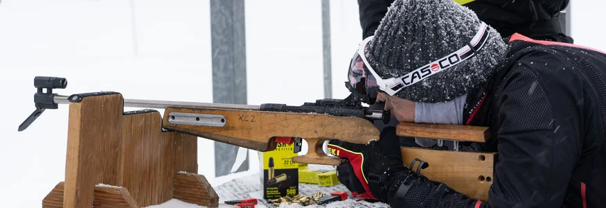 Person im Schnee zielt mit Gewehr auf einem Holztisch in liegender Position