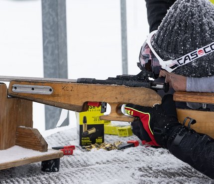 Person im Schnee zielt mit Gewehr auf einem Holztisch in liegender Position