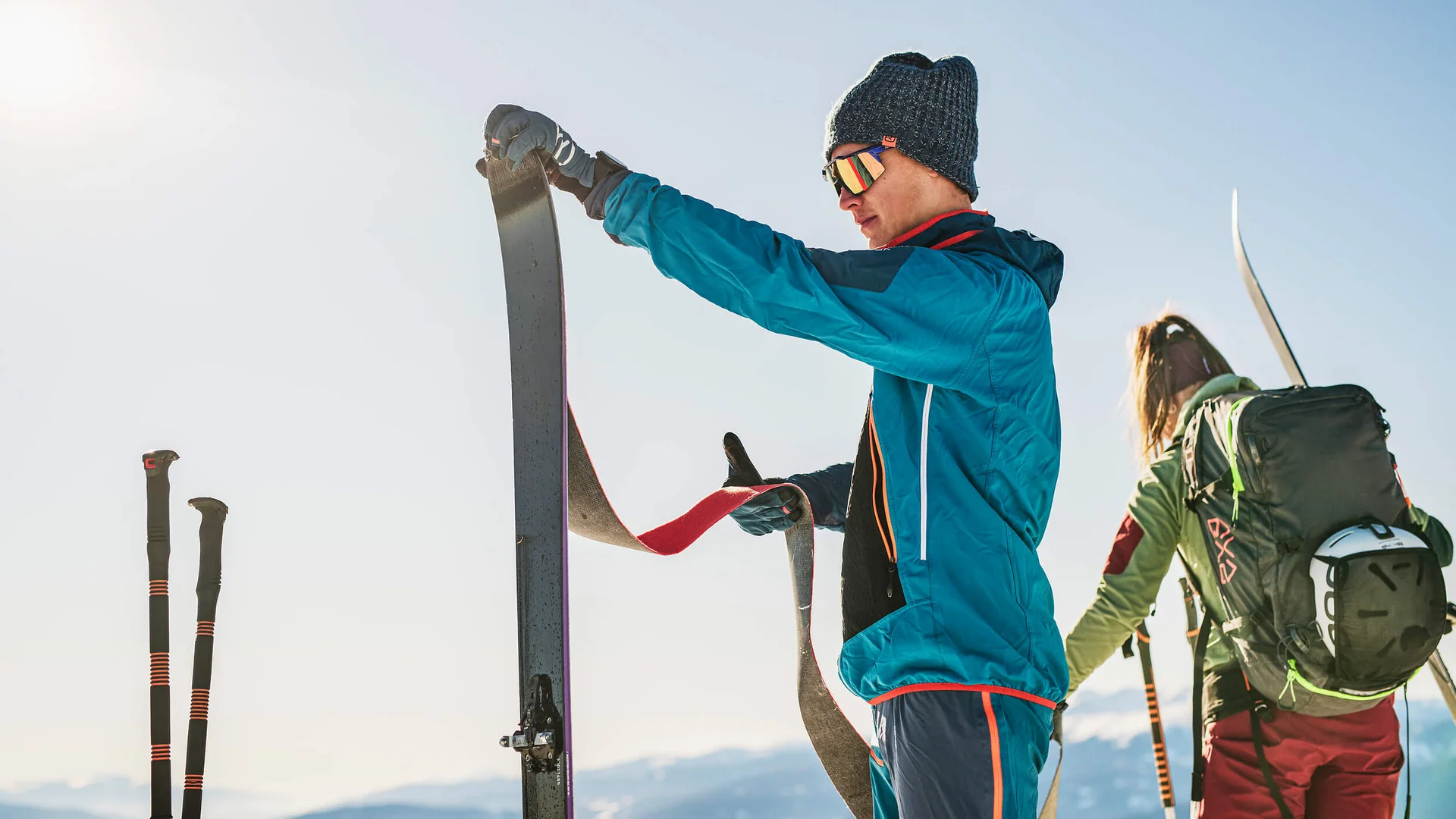 Mann bereitet Ski mit Fellen vor, Frau mit Rucksack im Hintergrund im Schnee