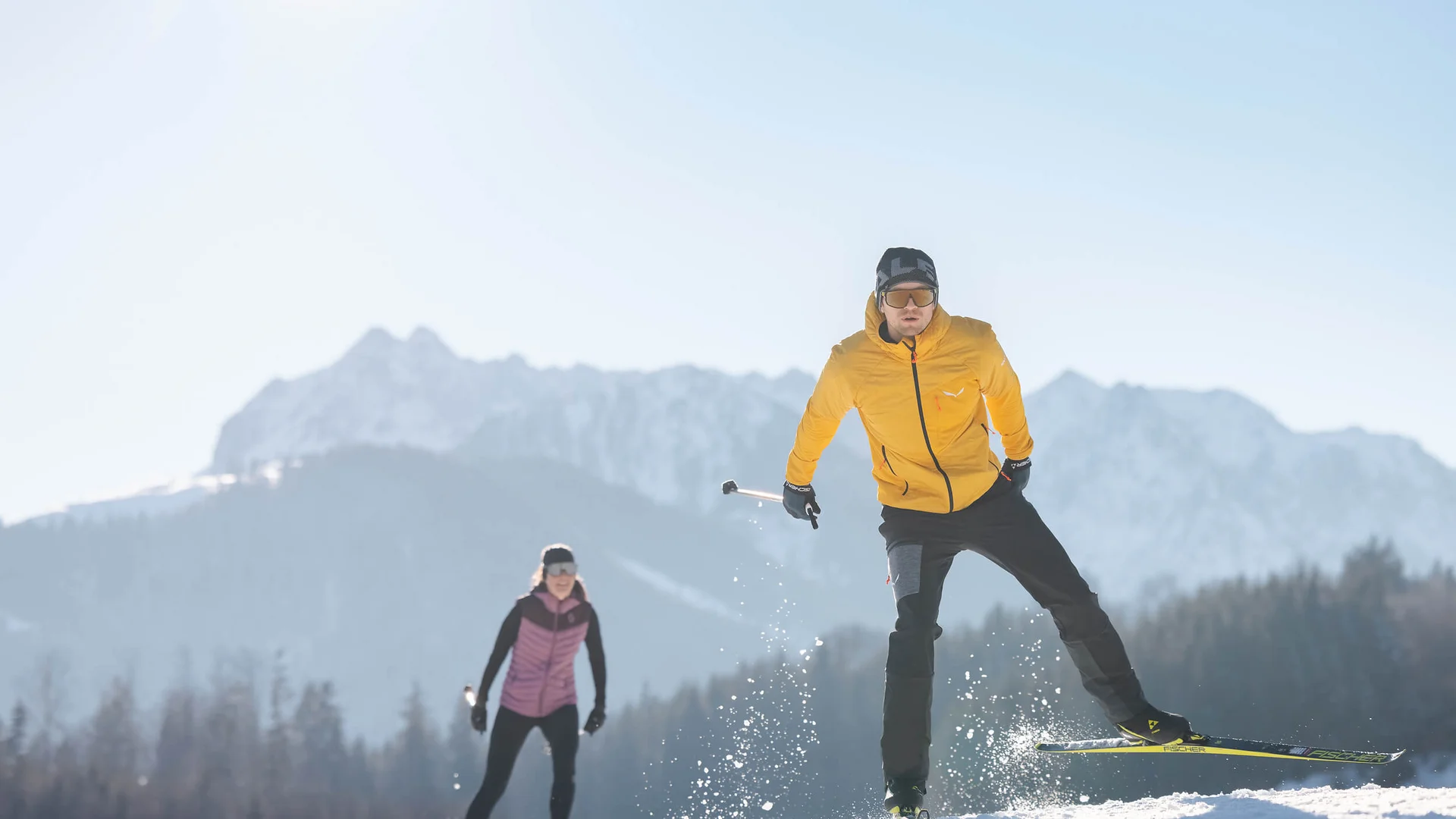 Zwei Langläufer auf schneebedecktem Feld vor Bergkulisse bei Sonnenschein