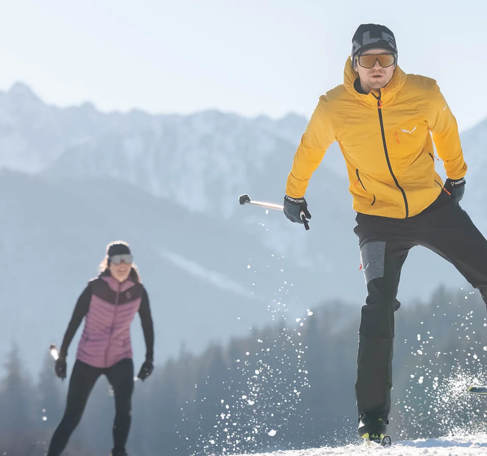 Zwei Langläufer auf schneebedecktem Feld vor Bergkulisse bei Sonnenschein