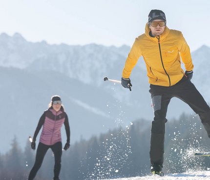 Zwei Langläufer auf schneebedecktem Feld vor Bergkulisse bei Sonnenschein