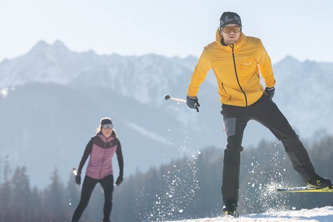 Zwei Langläufer auf schneebedecktem Feld vor Bergkulisse bei Sonnenschein