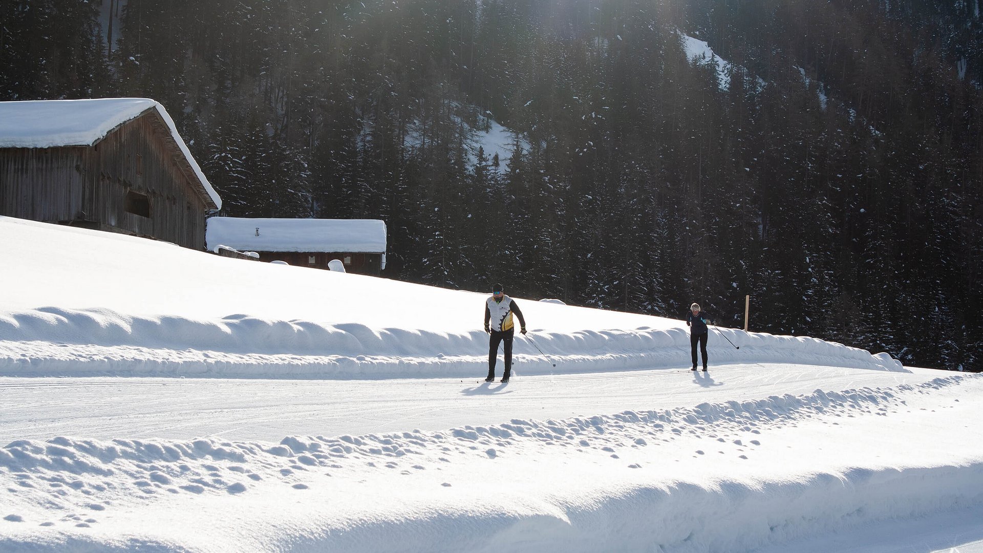 Zwei Langläufer auf verschneiter Strecke vor verschneiten Hütten und Wald