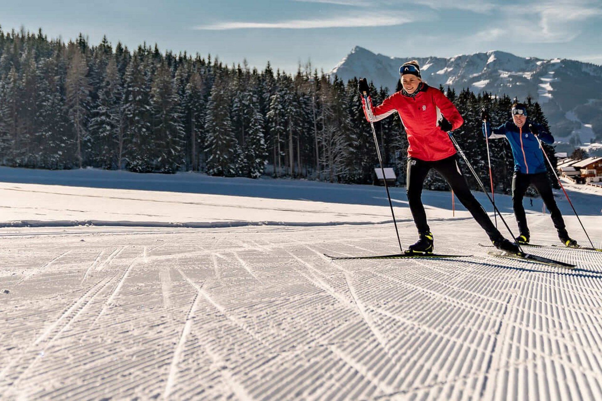 Two cross-country skiers on groomed trail in sunny winter mountain landscape