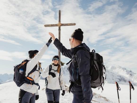 Three hikers giving a high five near a summit cross in snowy mountains
