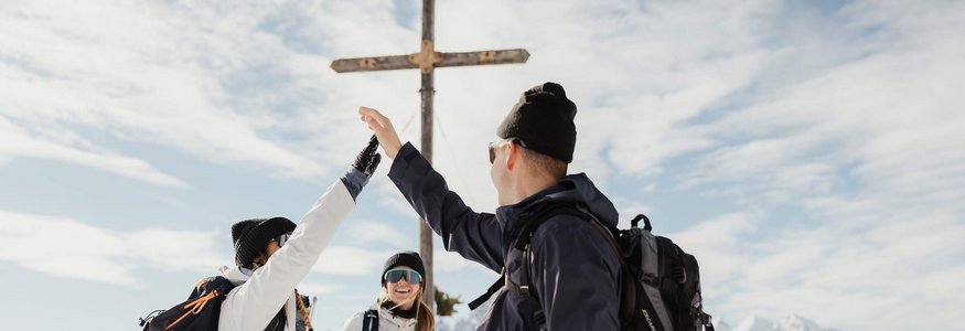 Drei Wanderer geben sich im Schnee an einem Gipfelkreuz einen High Five