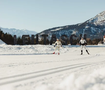 Two cross-country skiers in front of Leutasch sign with snowy mountains in background