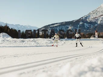 Two cross-country skiers in front of Leutasch sign with snowy mountains in background