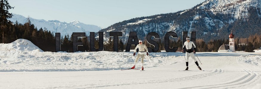 Two cross-country skiers in front of Leutasch sign with snowy mountains in background