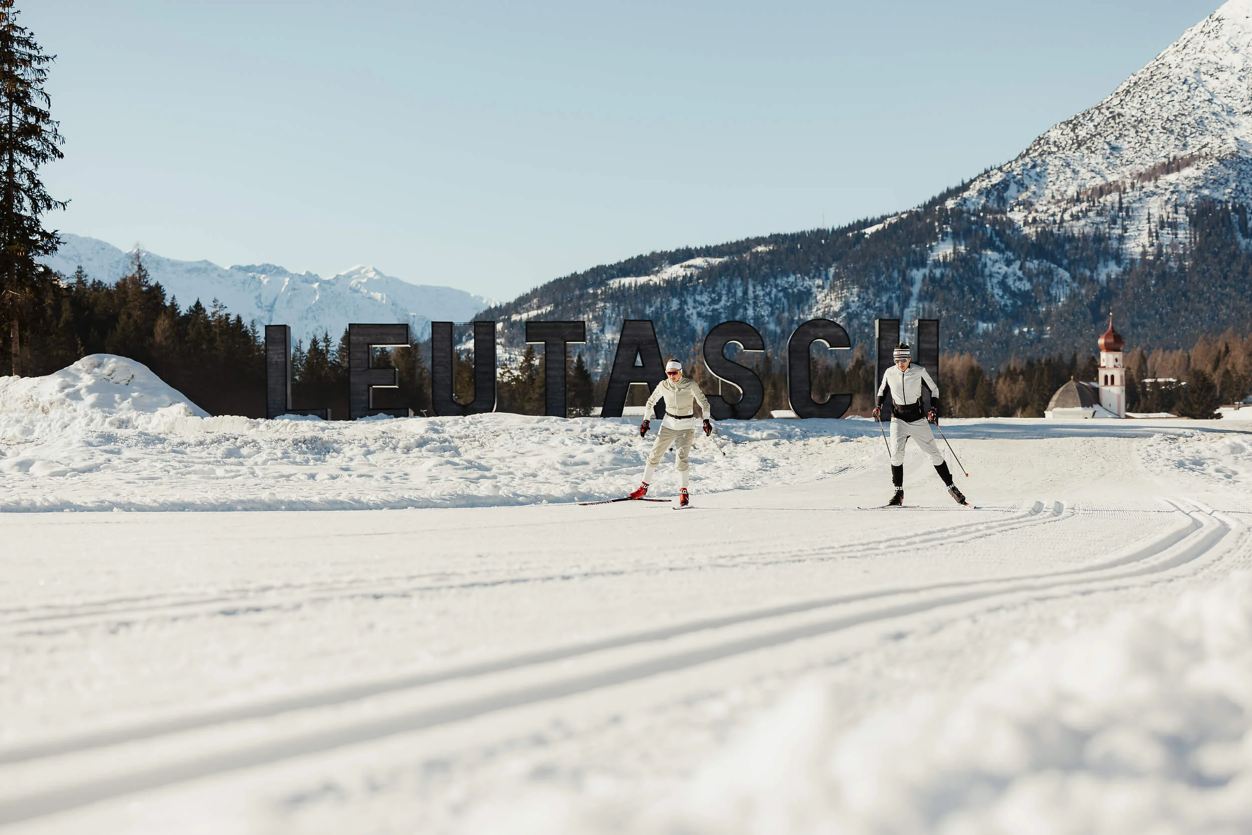 Zwei Skifahrer beim Langlauf vor Leutasch-Schrift im Schnee mit Bergen im Hintergrund