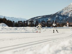 Two cross-country skiers in front of Leutasch sign with snowy mountains in background