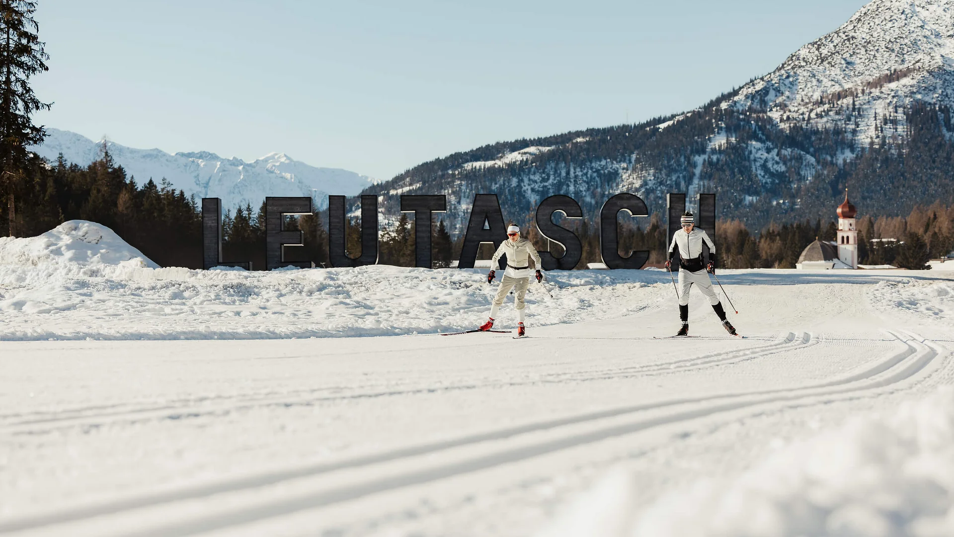 Zwei Skifahrer beim Langlauf vor Leutasch-Schrift im Schnee mit Bergen im Hintergrund