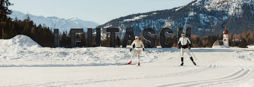 Zwei Skifahrer beim Langlauf vor Leutasch-Schrift im Schnee mit Bergen im Hintergrund