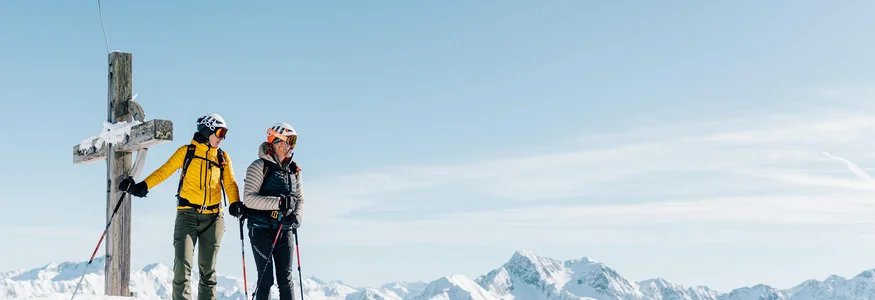 Zwei Skifahrer stehen am Gipfel neben einem Kreuz mit verschneiten Bergen im Hintergrund