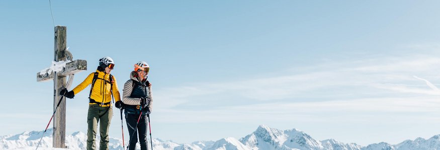 Zwei Skifahrer stehen am Gipfel neben einem Kreuz mit verschneiten Bergen im Hintergrund