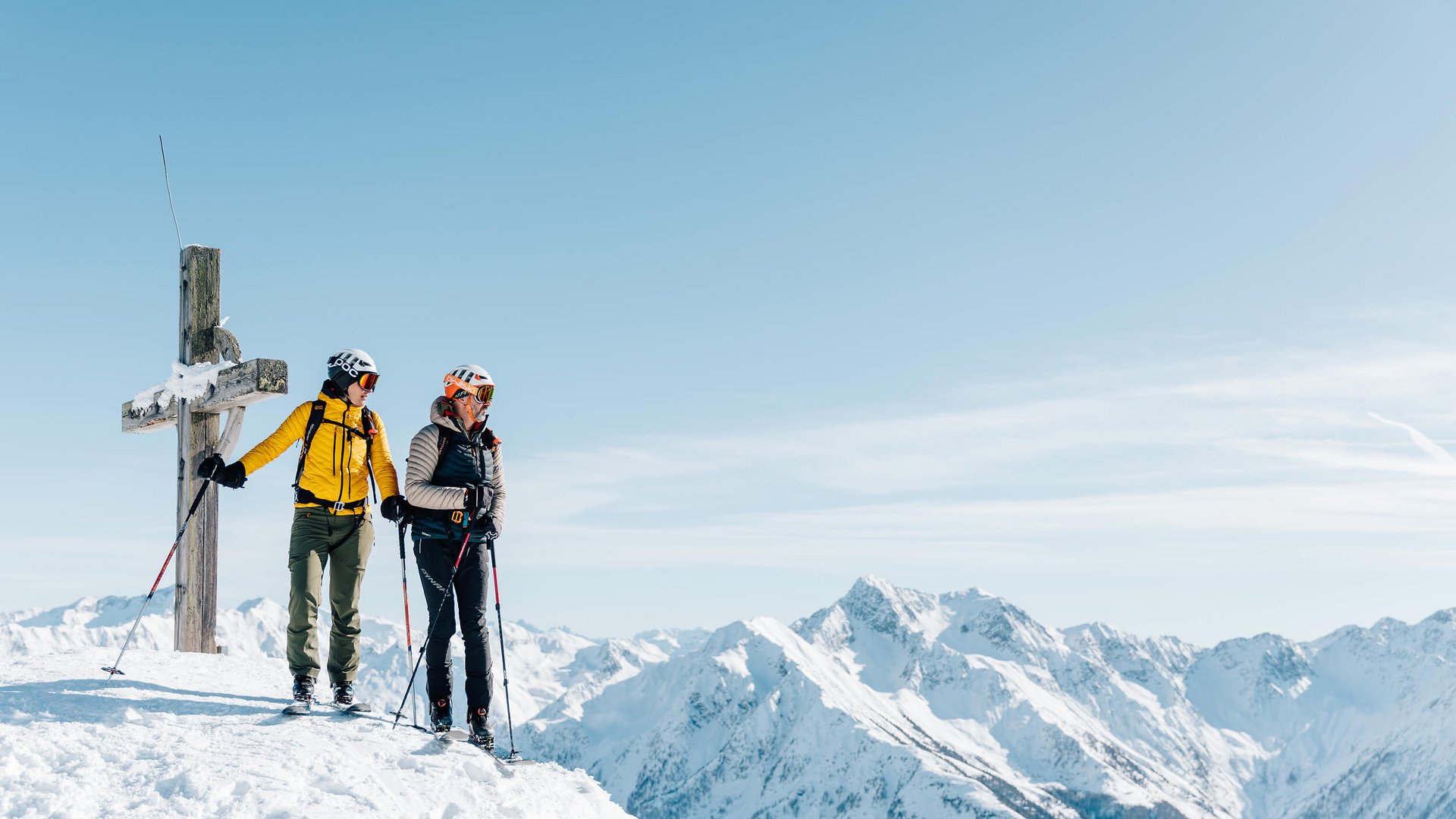 Zwei Skifahrer stehen am Gipfel neben einem Kreuz mit verschneiten Bergen im Hintergrund