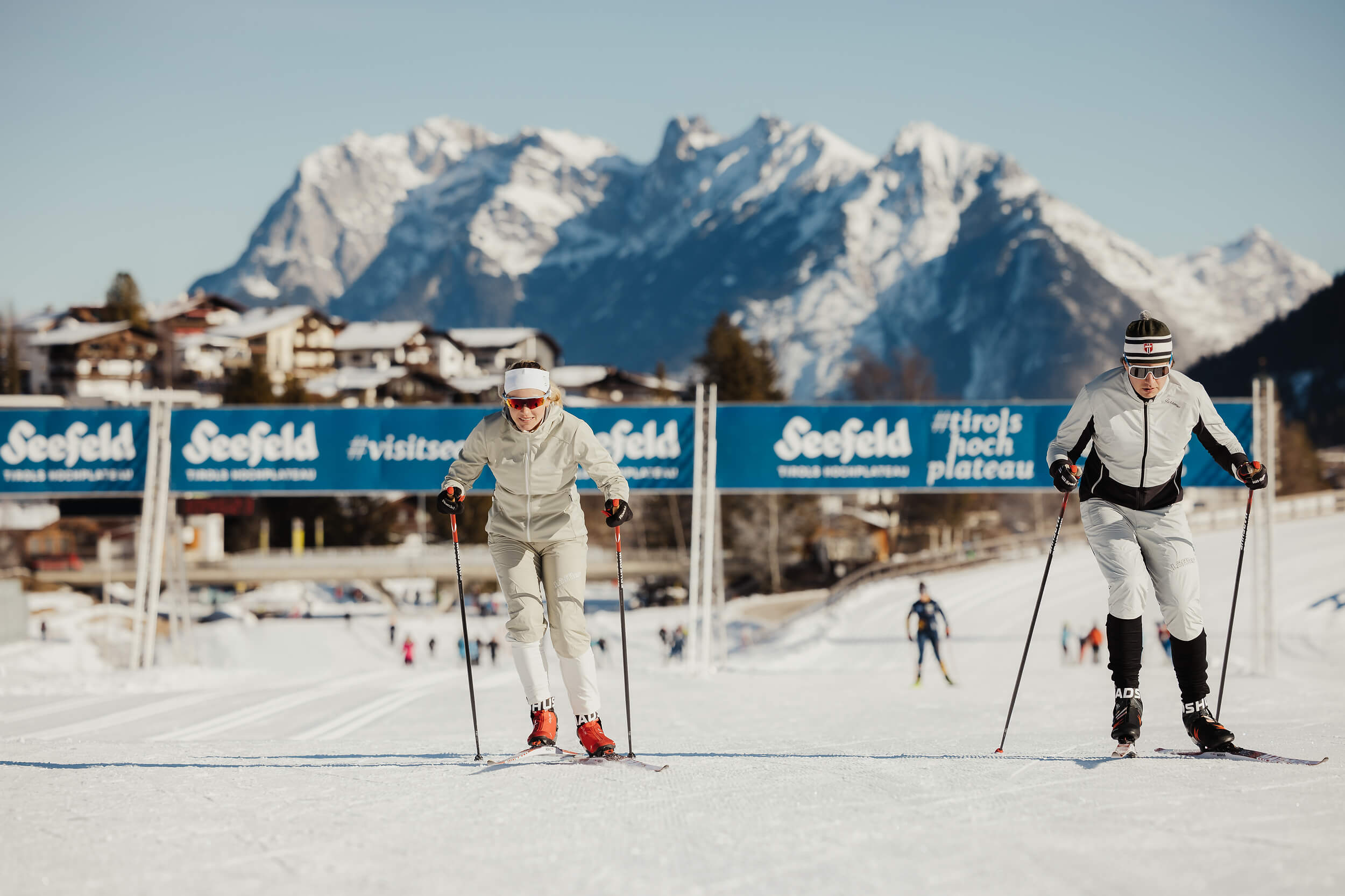 Zwei Langläufer auf der Loipe mit schneebedeckten Bergen im Hintergrund